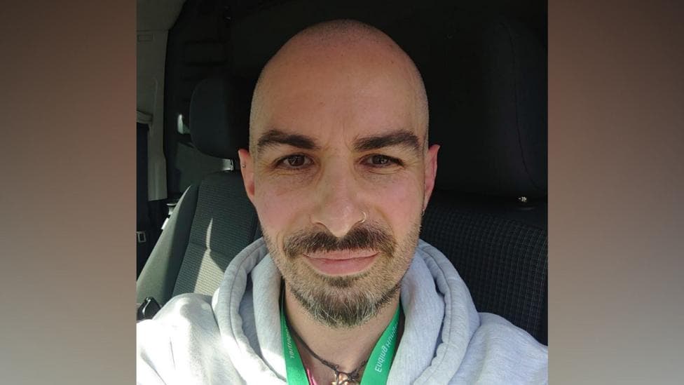 Man with short hair, beard and nose ring. He is wearing a grey hoody with a green lanyard. He is seated in a vehicle.