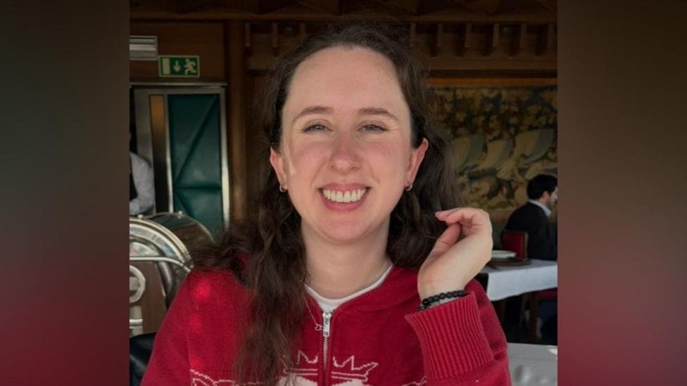 A young woman is smiling at the camera. She is wearing a red jumper with a zip up the front. She has long dark hair and pale skin.