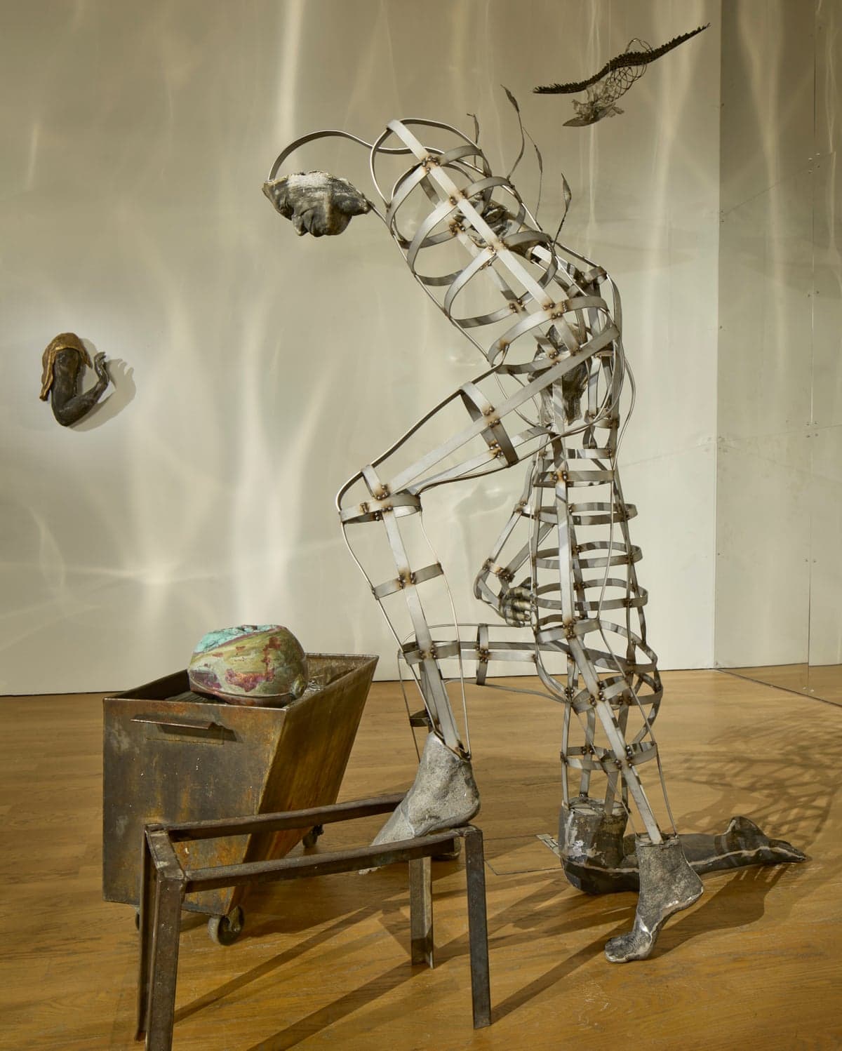 Kira Freije's wire-frame human sculpture bends forward beside a rusted cart, in a gallery