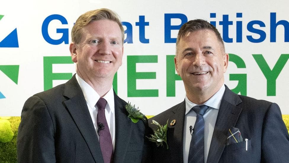 Two men in dark suits stand in front of a sign which says Great British Energy