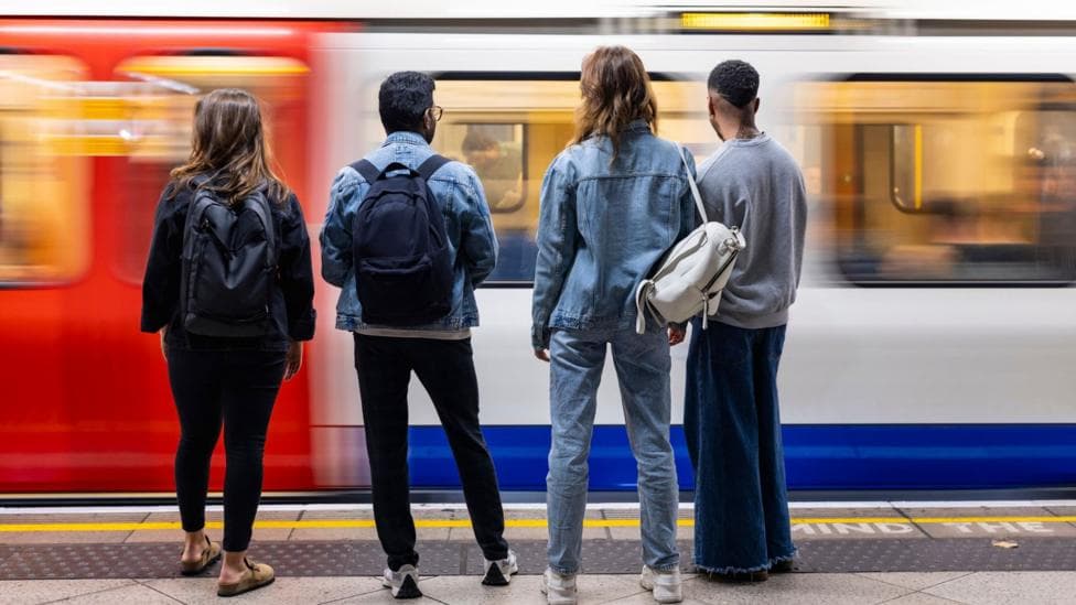 Four young people stand on a London underground platform facing the tracks as a train arrives.