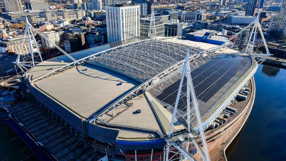 The Principality Stadium, a large stadium with four spires on each of its four corners, towers over the city of Cardiff. The picture is a birds eye view picture taken from above the stadium. The photo is taken on a sunny day.