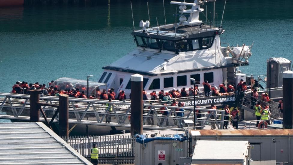 A vessel at a quayside with "border security command" written on the hull. Dozens of people wearing life jackets are filing off the boat and onto a pontoon.