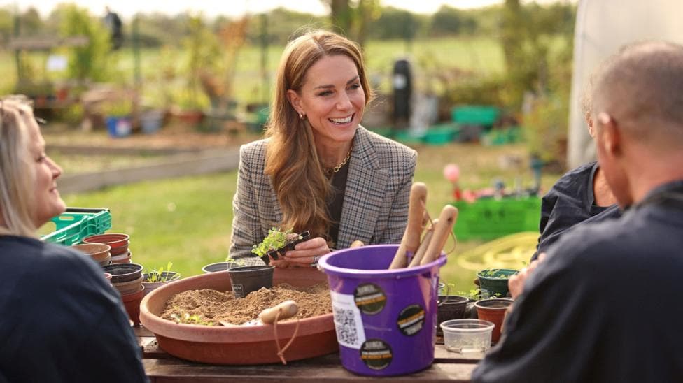Catherine, princess of Wales, dressed in a grey and blue Tweed-style jacket and dark top smiles in the centre of the image. She has long brown hair. She is sitting at an outdoor round table, on which are plastic gardening pots and hand tools in a purple bucket. A man with a shaved head to the right, and a blonde-haired woman, to the left, are seated at the same table, their backs to the camera. Both are wearing dark clothing.