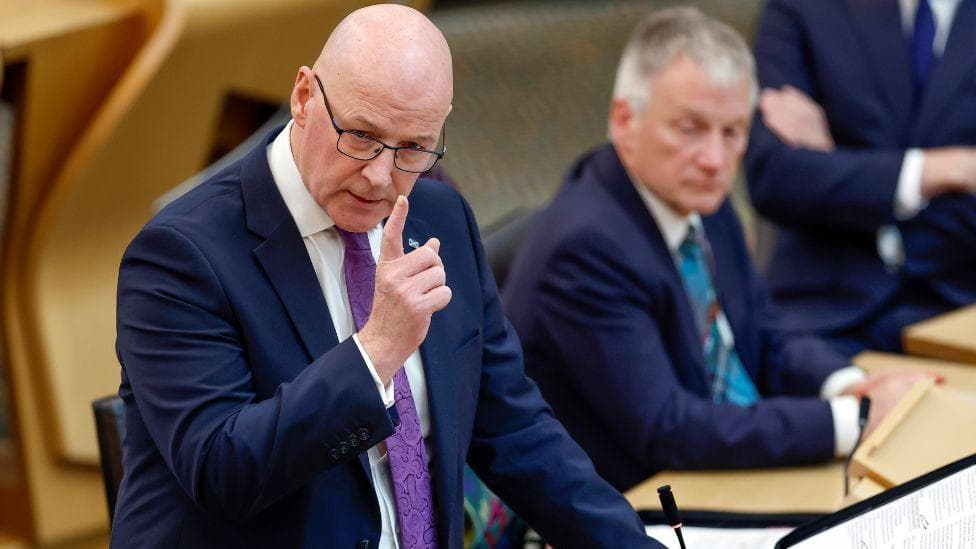 John Swinney, who is bald with glasses, speaks with his right hand raised in the parliament chamber