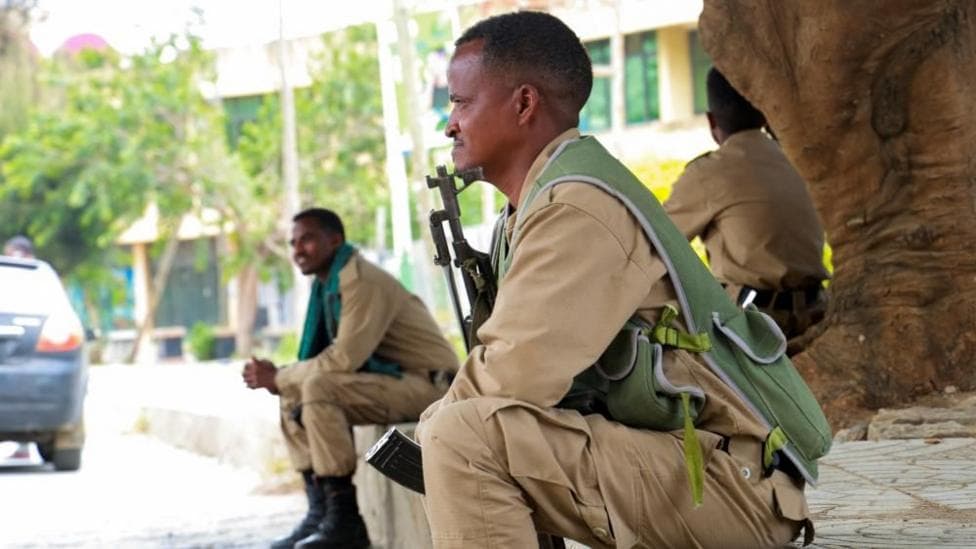 A soldier in khaki seen in profile. He is sitting on a stone step, holding am AK47 between his legs. Out of focus, another soldier can be seen behind him.