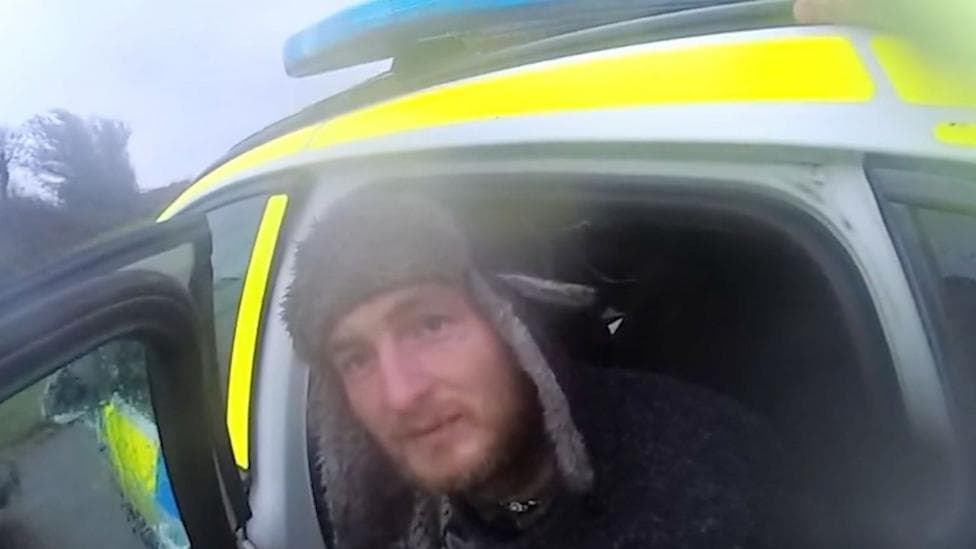 A grab from police body worn camera footage showing Alexander Lewis-Ranwell sitting with his head out of the back door of a police car. The yellow and blue livery of the police vehicle is partially visible. Lewis-Ranwell is looking towards the camera and is wearing a dark coloured hat with light coloured fleece covering the forehead area and ear flaps.
