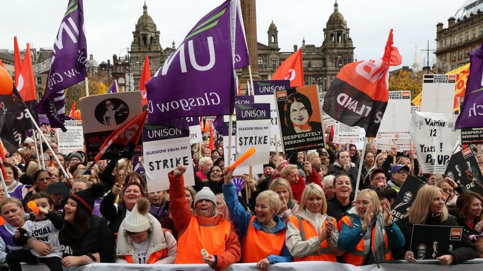 A large group of women stand behind a banner that reads 'Glasgow Equal Pay'. They are wearing orange high visibility vests and standing with their arms raised, shouting and waving Unison flags. In the background is the Glasgow City Council building in George Square.