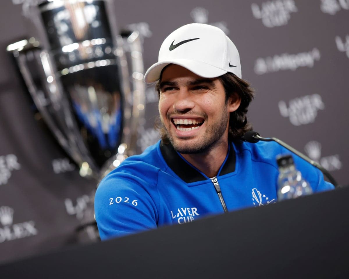 Carlos Alcaraz, wearing a Laver Cup branded jacket and a Nike hat and sitting in front of the trophy, laughs during a press conference to promote the 2026 Laver Cup in London.