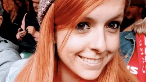 Danielle is shown smiling and wearing a beanie hat in a crowd. Her hair is ginger coloured and she is looking at the camera with her brown eyes.