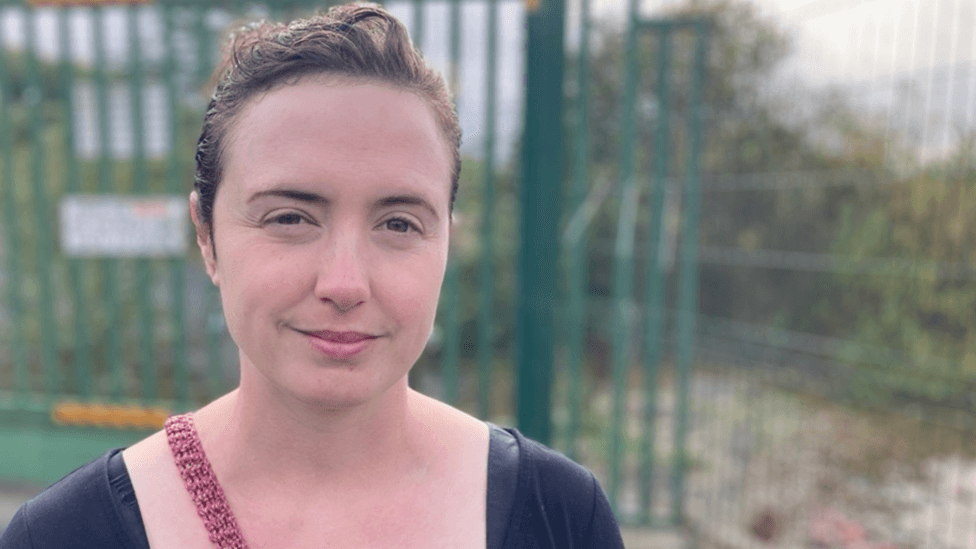 Natalie Dugdale is a white lady with brown hair wearing a blue t-shirt and a red handbag in front of a wooded area