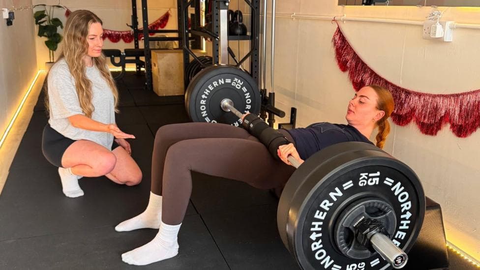 Antonia, on the left, wearing a grey t-shirt and black shorts, is talking to a young female client while she is doing hip thrusts using heavy weights on a bar which is lying across her hips