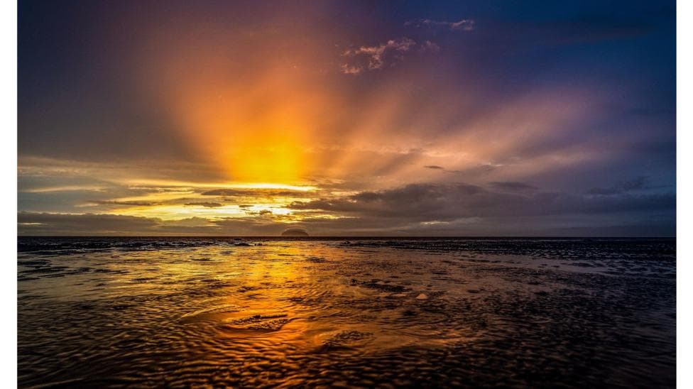 Golden sunlight breaks through dramatic clouds at the horizon, casting vibrant orange and amber reflections across a wet tidal shoreline. Dark storm clouds and deep blue tones contrast with the warm glow of the setting sun, creating a striking and atmospheric sky. The rippled sand and shallow water in the foreground mirror the colours above, adding depth and texture to the coastal scene.