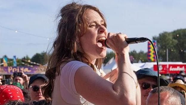 Kate Nash pictured in a white top singing into a handheld microphone while standing among a large crowd at an outdoor music festival. Audience members surround the performer, with some filming on smartphones. Colourful festival banners, food stalls and a line of trees are visible in the background under a bright blue sky.