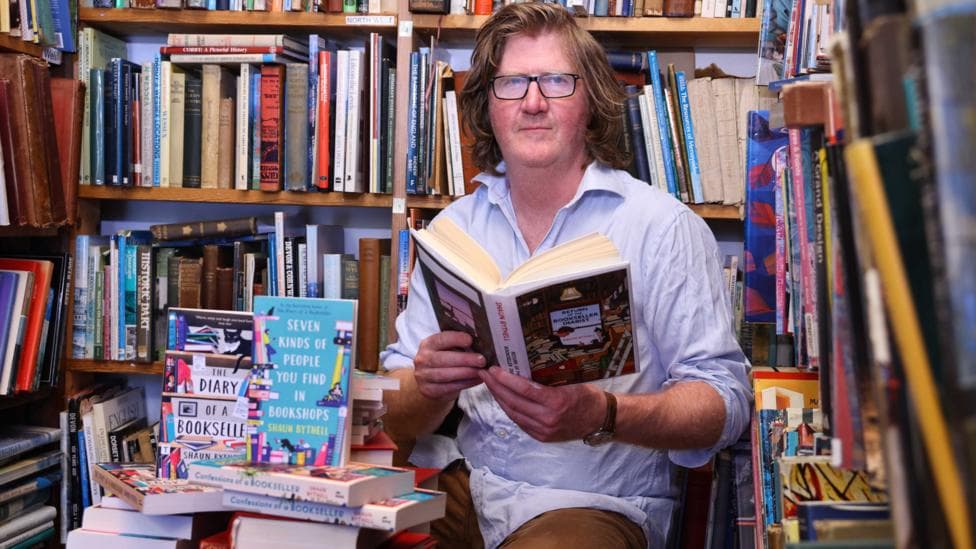 A man with long reddish brown hair in a blue shirt sits reading a book surrounded by books
