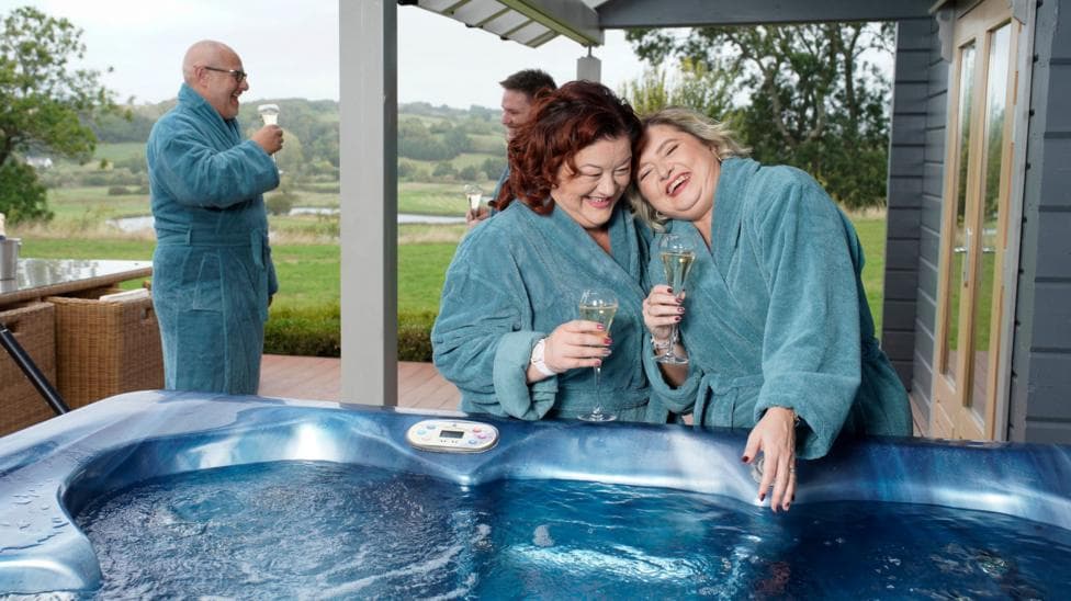 Amanda and Louise in blue bathrobes holding a glass of champagne each and standing by a hot tub.
