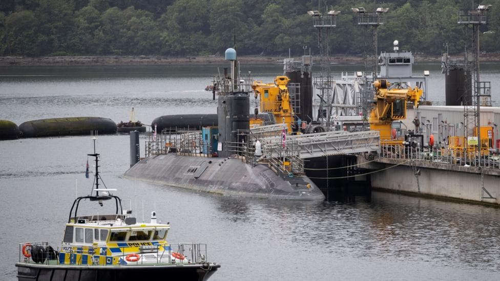 A large dark-grey submarine is moored alongside a naval dock on a calm body of water.
Industrial equipment, cranes, gangways and support structures line the pier, with a police boat nearby.