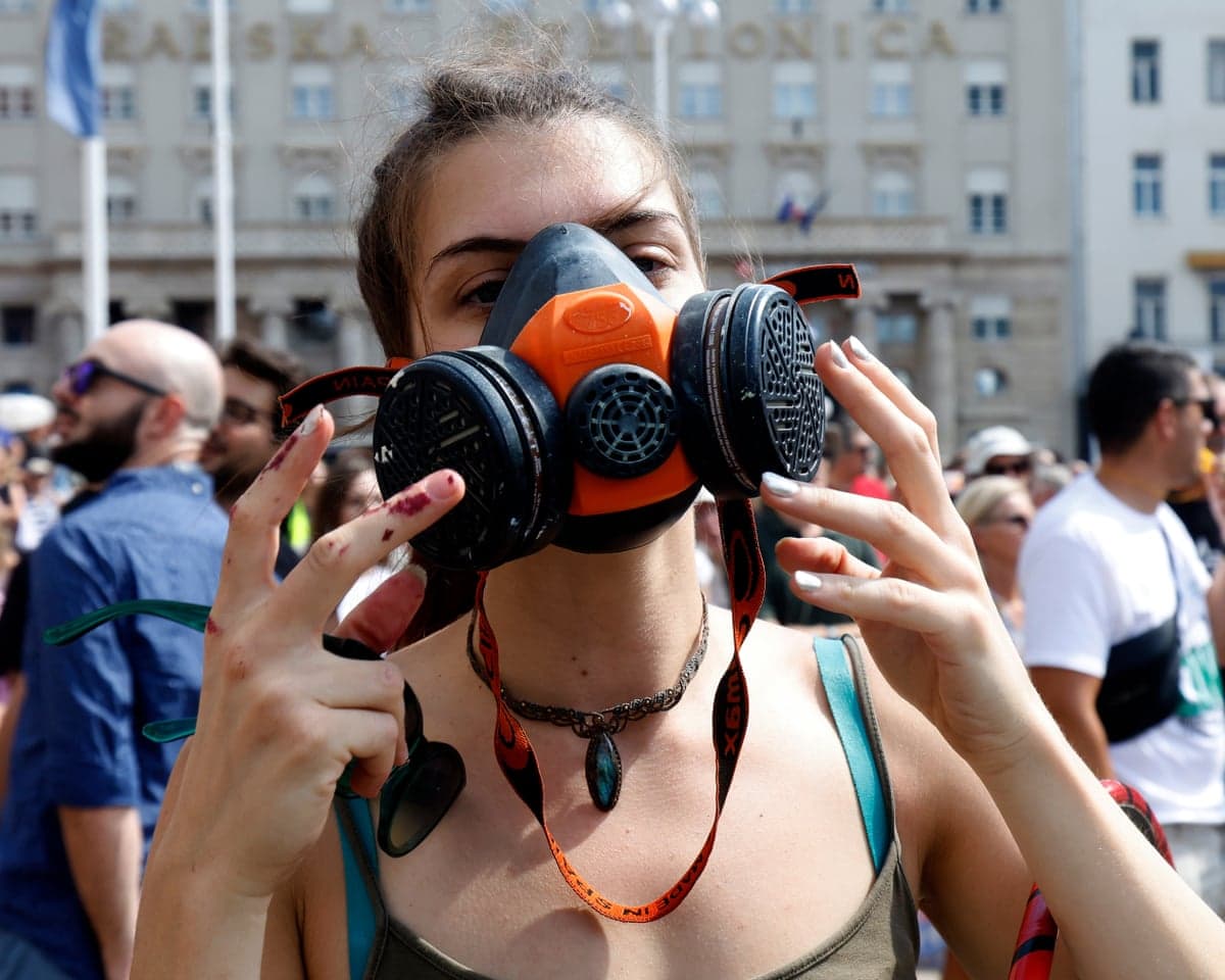 A woman wearing a gas mask raises her hands to her face, with fellow protesters visible in the background.