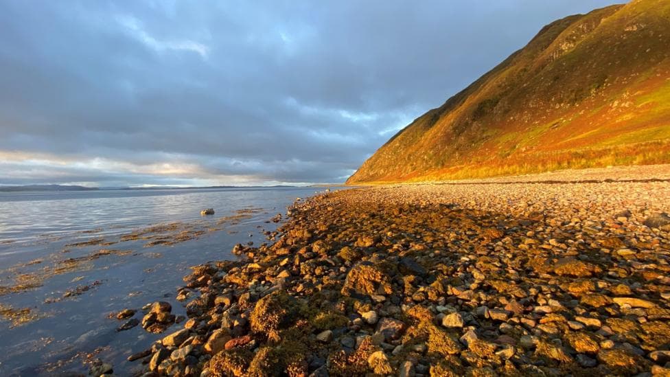 Rocky shoreline and calm sea beneath a sunlit hillside, with dramatic dark clouds overhead.