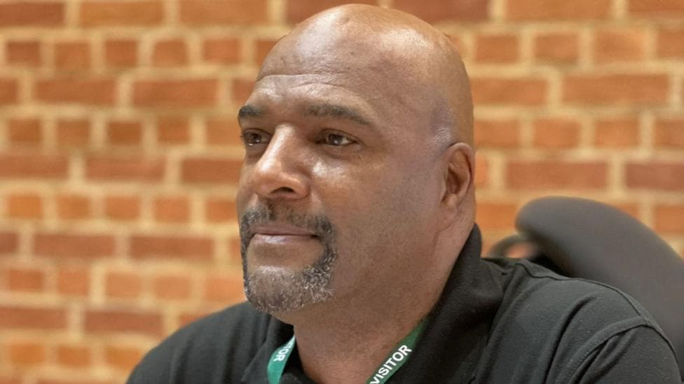 A man with short facial hair, wearing a black shirt and a green lanyard reading "visitor". He is pictured from the shoulders up and there is a brick wall behind him.
