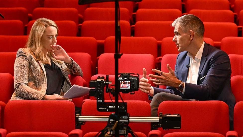 Laura Kuenssberg and Matt Brittin sitting in read theatre-style seats and speaking during the interview, with a camera filming them