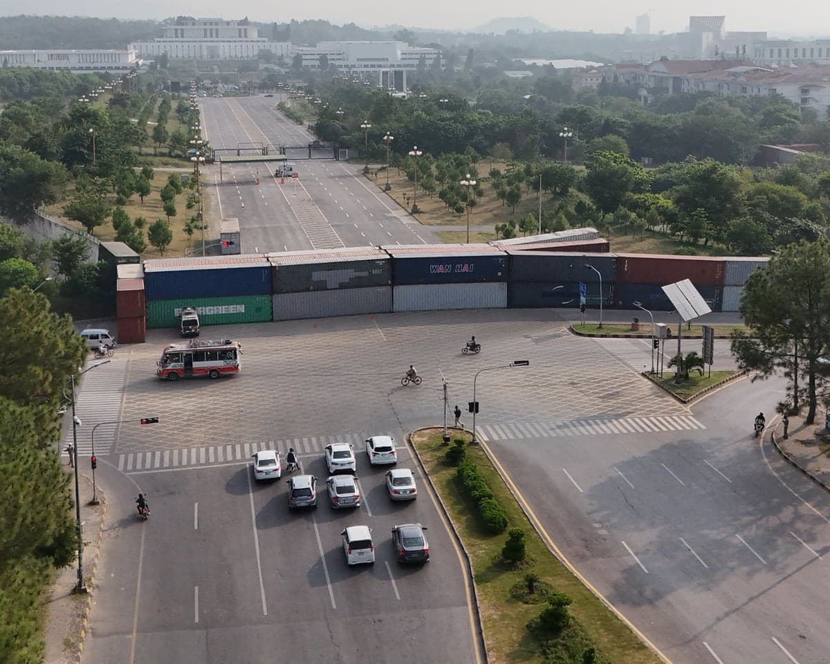 An aerial shot of shipping containers stacked across a road, with traffic held up in the foreground