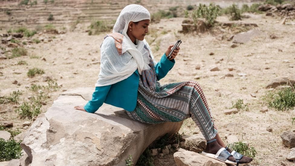 A woman sits on a rock in an arid landscape looking at her phone. She is wearing a white headscarf, blue jacket and a patterned skirt