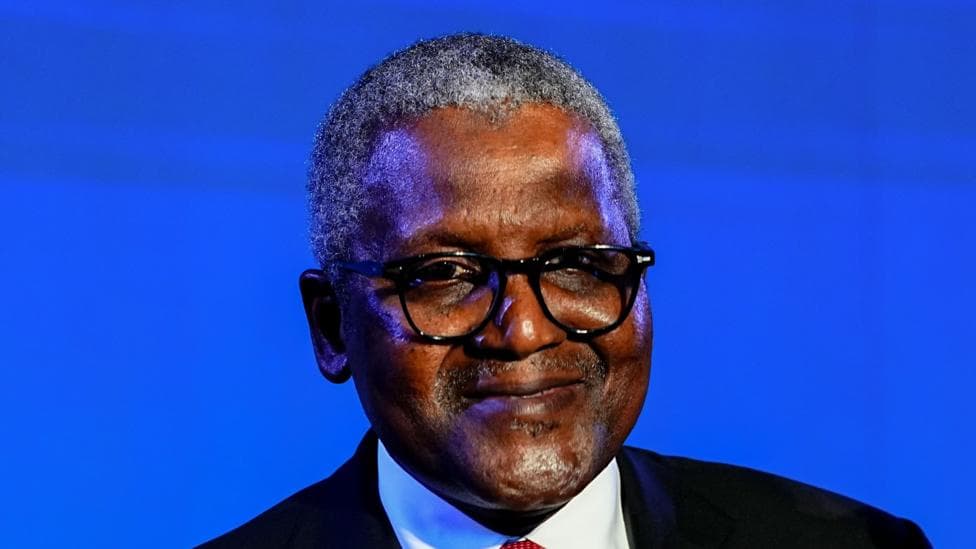 A man with grey hair, wearing glasses with black frames, a black suit, a white shirt, and a red tie, smiles into the camera