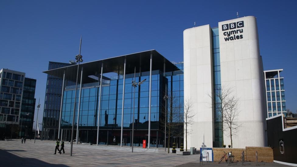 A huge glass building with pillars on central square. The white brick has black letters on it saying 'BBC Cymru Wales'. The background is a blue sky.