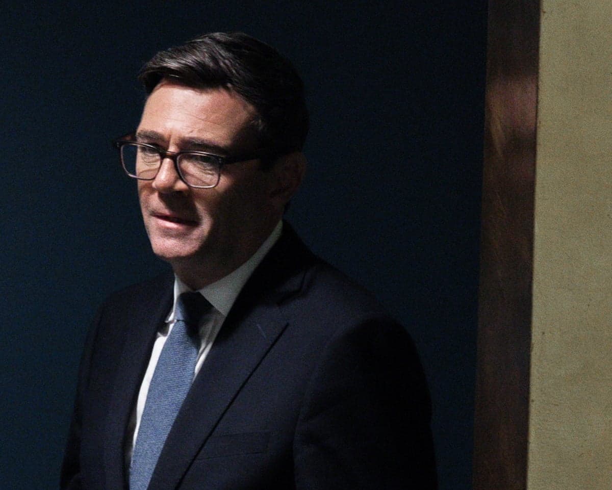 Andy Burnham, in glasses and a dark suit, walks into a dimly lit room, looking downward
