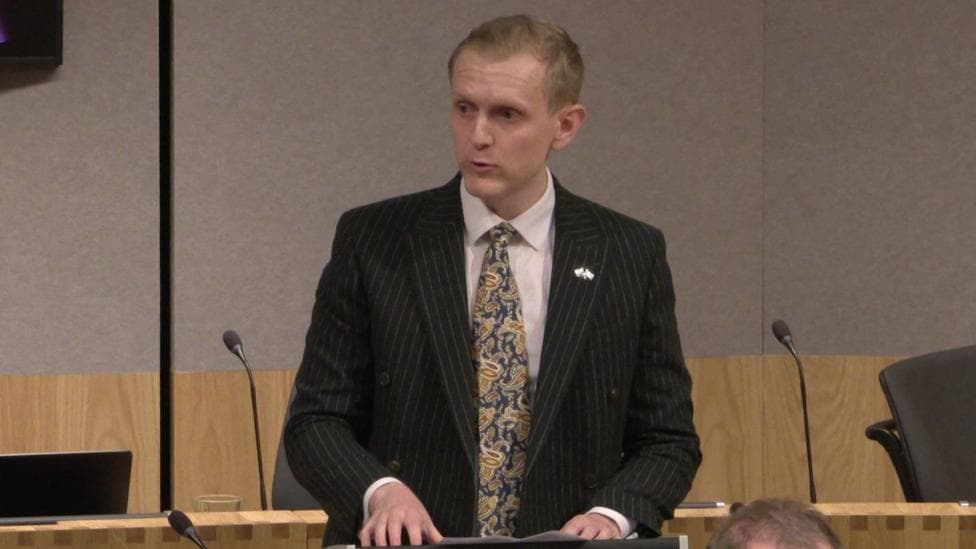 Joe Martin, holding his hands over a lecturn which has a piece of paper visible on it. He appears to be speaking to the left hand side of the photograph, and is wearing a striped suit and patterned tie.