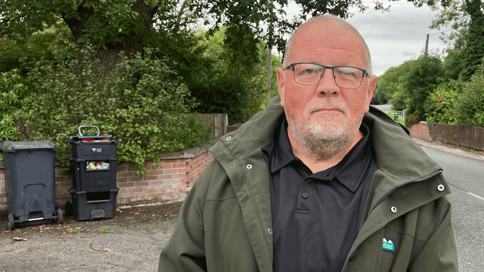 Alan Jones is wearing a green jacket and black shirt. He is standing on roadside flanked by green trees and bushes. There are recycling bins against a brick wall
