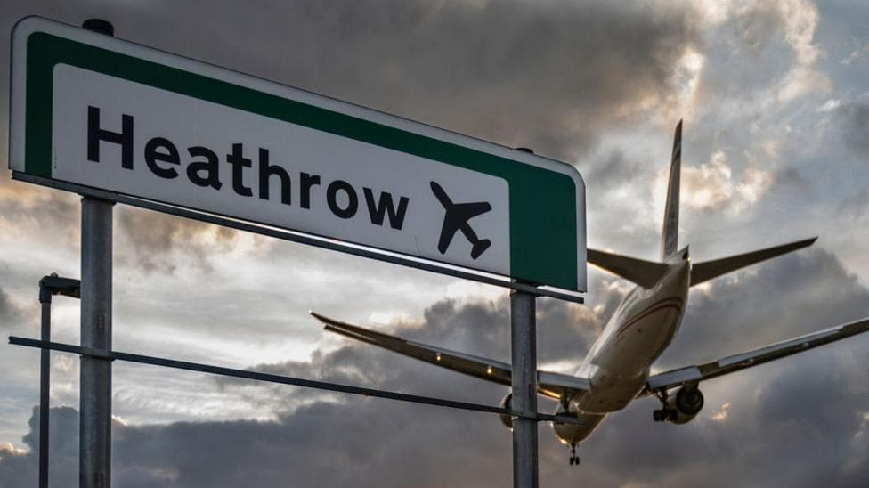 A commercial aeroplane flying low on final approach to land at Heathrow Airport, passing directly over an overhead highway road sign.