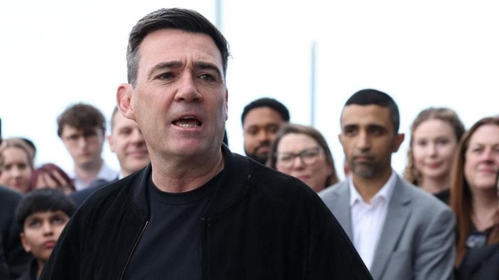 Andy Burnham is wearing a dark jacket over a dark top. He is stood in front of a crowd of people and is speaking.