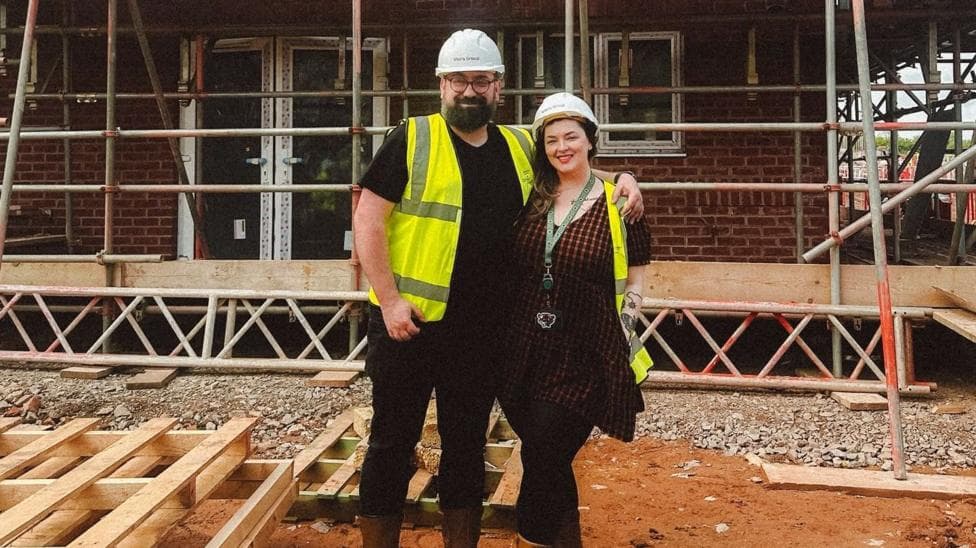 Charles and Georgina pose on a muddy building site in front of a partially completed brick house surrounded by scaffolding. Both wear safety helmets, high-visibility vests and sturdy boots. Stacks of timber lie in the foreground, while the unfinished structure, exposed windows and metal scaffold framework fill the background, illustrating the progress of the construction project.