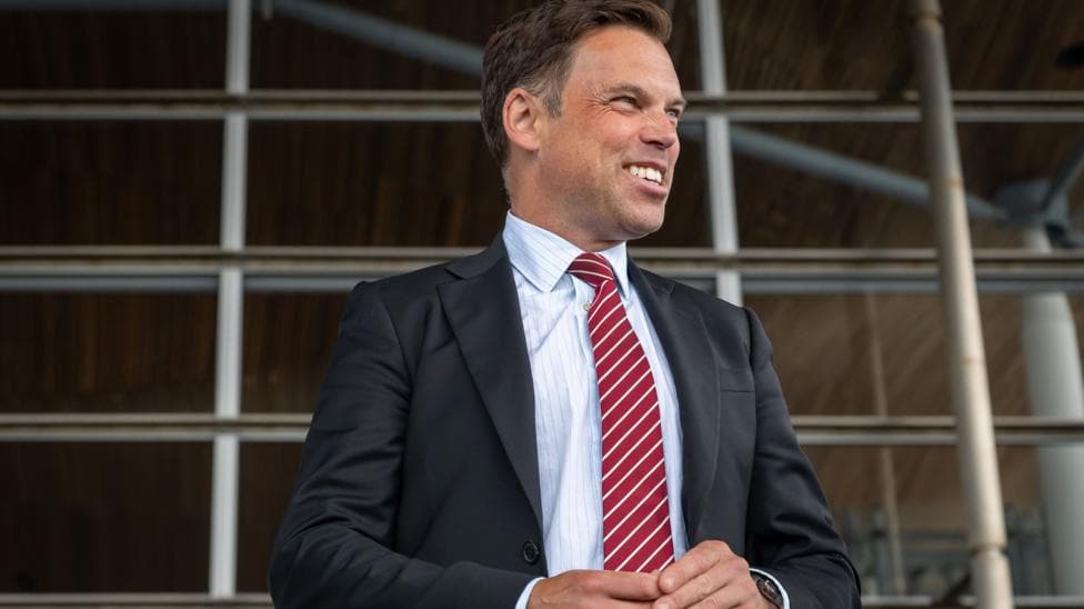 The image shows Ken Skates standing outside the Senedd. He is smiling and looking to his left. He is wearing a dark suit, white shirt and stripey red and white tie.