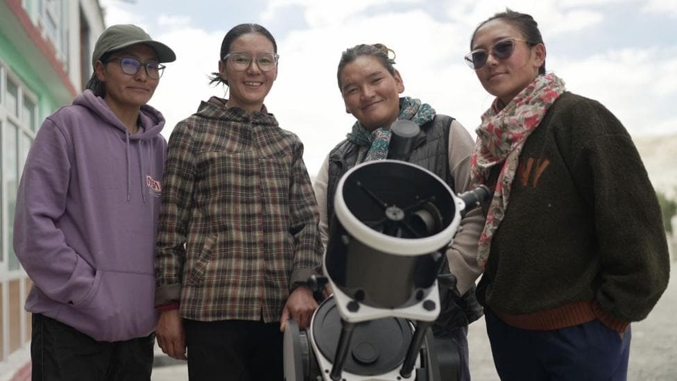 Padma Chamchot, Tsering Yangdol, Tsering Dolker and Palzes Angmo stand outdoors around a large telescope, looking toward the camera. The telescope is positioned in the foreground, with a building on the left and a cloudy sky in the background. The group is dressed in casual jackets, hoodies, and scarves, suggesting a community or educational astronomy activity.