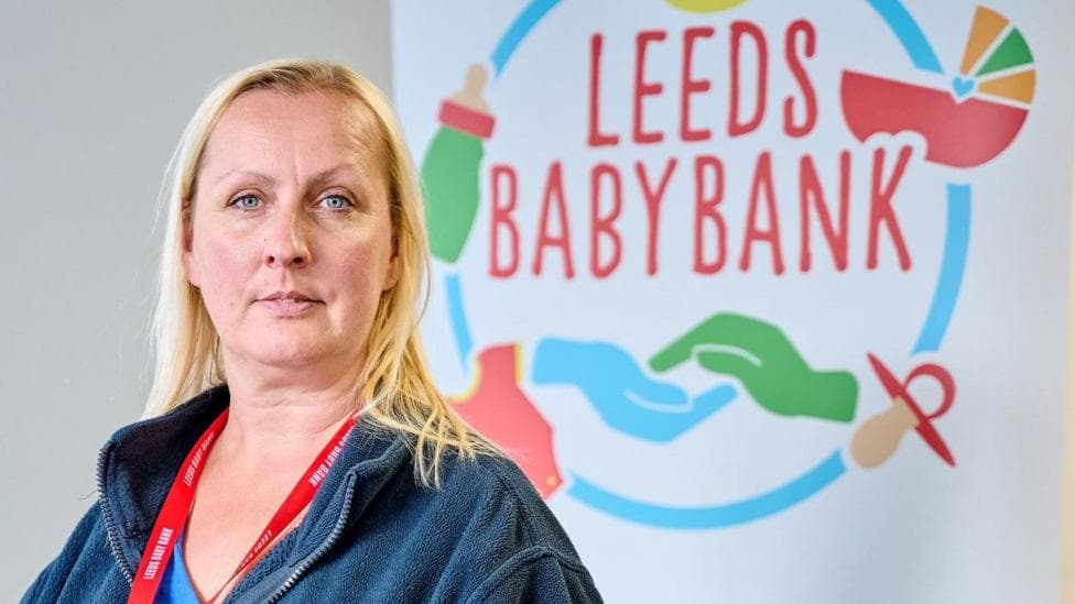 A woman with shoulder-length blonde hair and blue eyes looks into the camera with a neutral expression. She is wearing a dark blue fleece jacket and has a red lanyard around her neck. In the background is a large sign with Leeds Baby Bank on it, along with images of hands, dummies, prams and baby bottles