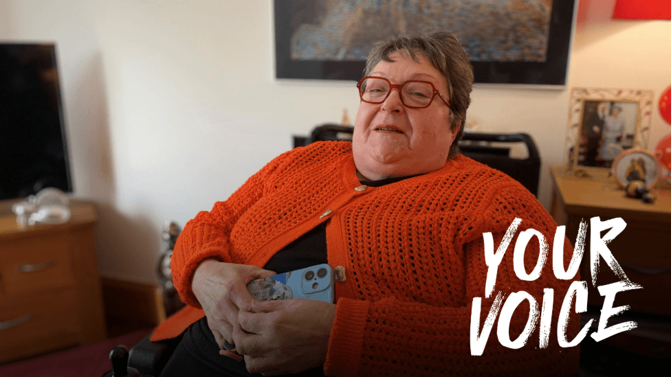 A woman in a wheelchair in a bright orange knitted cardigan. She is wearing glasses and is holding her mobile phone in her hands. She is sitting in her living room.