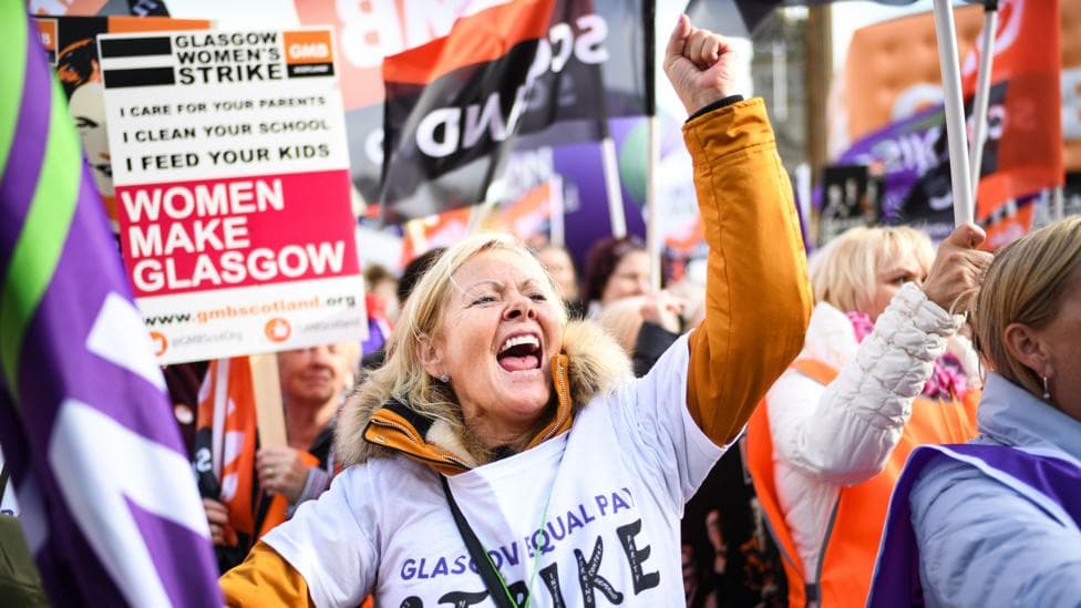 Protestors on a march. One woman is in the centre of the picture, punching the air and yelling. Many of the women around her are holding flags or placards, including one that says Glasgow Women's Strike and Women Make Glasgow.