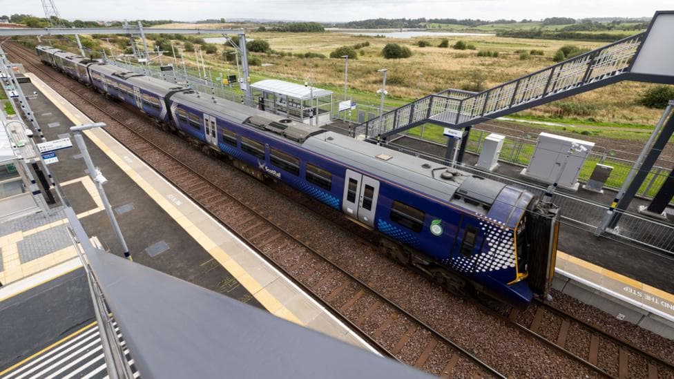 An overhead view of the new Balgray train station. It has two platforms and two tracks, with a blue train going along one of the tracks. Stairs with handrails are visible on each platform, as is a small waiting area with a roof.
Grassy fields are in the distance behind the station.