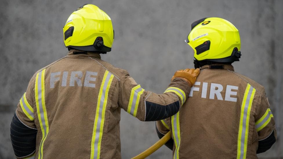 Two firefighters clad in fluorescent helmets and uniforms have their backs to the camera. One firefighter has their hand on the other's shoulder and a length of yellow hosepipe is visible in the middle.