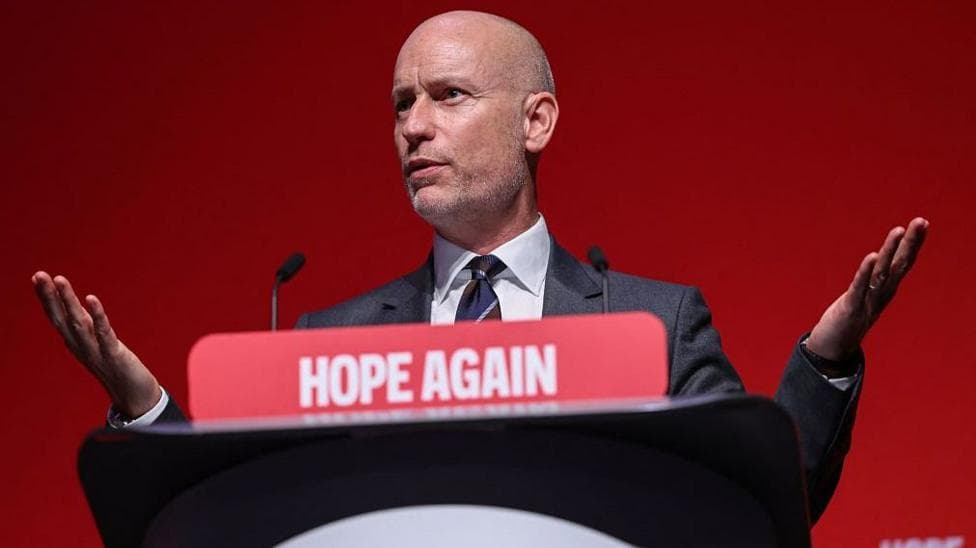 A bald man in a suit and tie, with his arms up in a frustrated gesture, stood at a podium and speaking into a microphone. On the front of the podium is a red sign with white lettters reading: HOPE AGAIN.