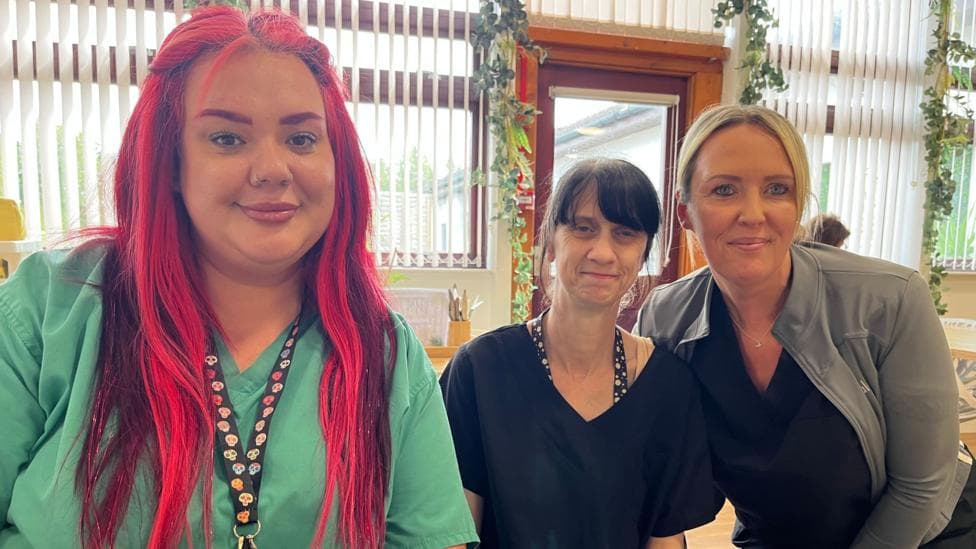 Three women sitting in a cafe. The woman on the left has long, pink hair and wears green scrubs The woman in the middle has her hair tied back and wears a black top. The woman on the right is in a black top and a grey jacket.