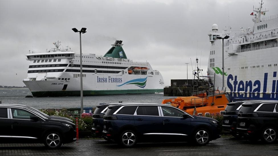 A port where the Irish Ferries boat and Stena Line are visible. They are white boats. One with Irish Ferries written in blue text. There is also green paint on the ferry. The Stena Line boat, which is to the right of the image, has blue text saying Stena Line. The boat is white. There are dark coloured cars parked at the port.