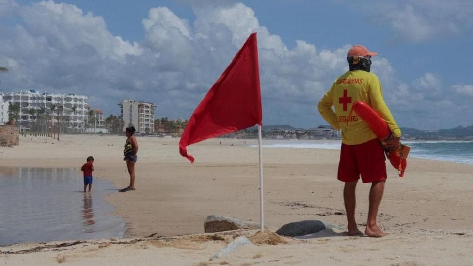 A lifeguard stands next to a red flag warning of hazardous sea conditions as Hurricane Polo approaches Baja California Sur in San Jose del Cabo, Mexico, on 26 September, 2026. The lifeguard is wearing a bright yellow long-sleeved shirt with the words "salvavidas" (Spanish for lifeguard) and "lifeguard" and a red cross on the back.