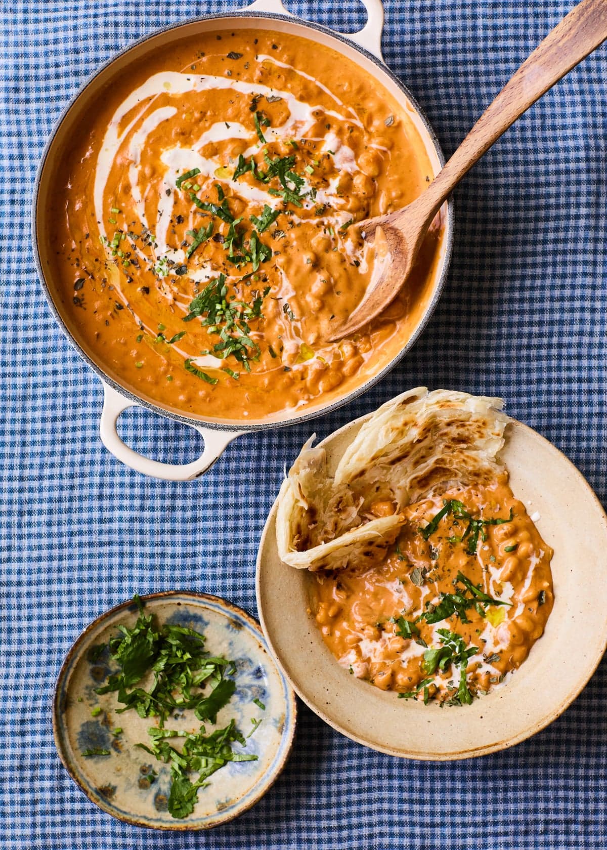 A bowl and a large pot of of baked beans butter masala with a bowl of chopped leaves on the side, all on a blue and white chequered cloth.