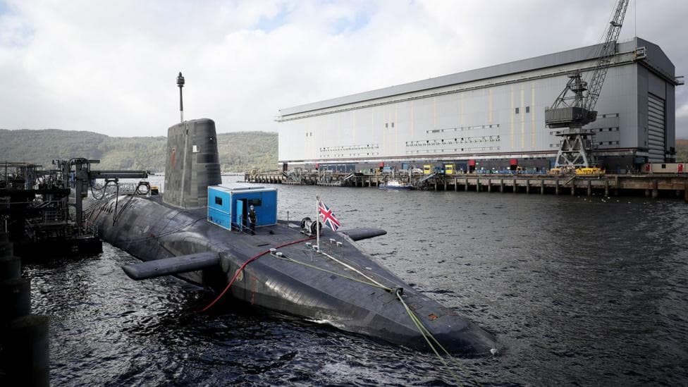 A submarine with a union flag flying from it, with a large white maintenance building in the background