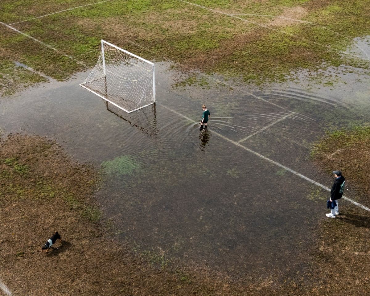 Two people and a dog standing in a flooded football pitch.