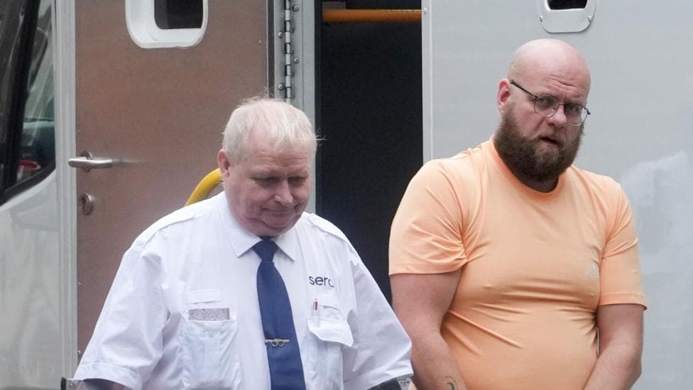 A man wearing an orange t-shirt. He is being led from a prison van by a custody officer.